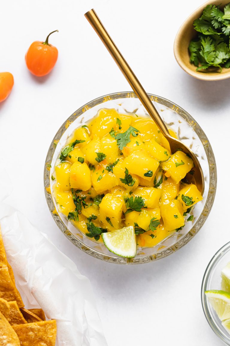 Mango habanero salsa in a bowl with a spoon, surrounded by cilantro, habanero peppers, limes, and chips.