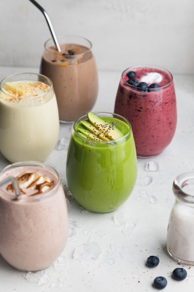 all 5 smoothies in cups with ice, blueberries, and almond milk on the side