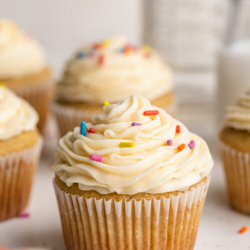 close up image of a gluten-free vanilla cupcake with vanilla frosting and rainbow sprinkles.