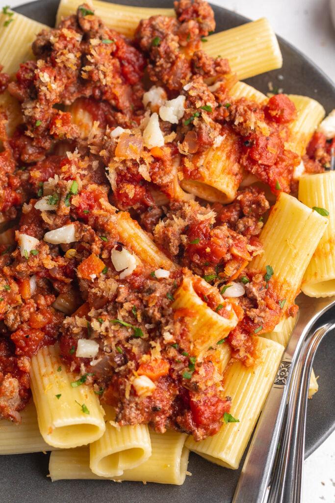 close-up image of rigatoni bolognese with parmesan cheese, toasted breadcrumbs, and chopped parsley on top.