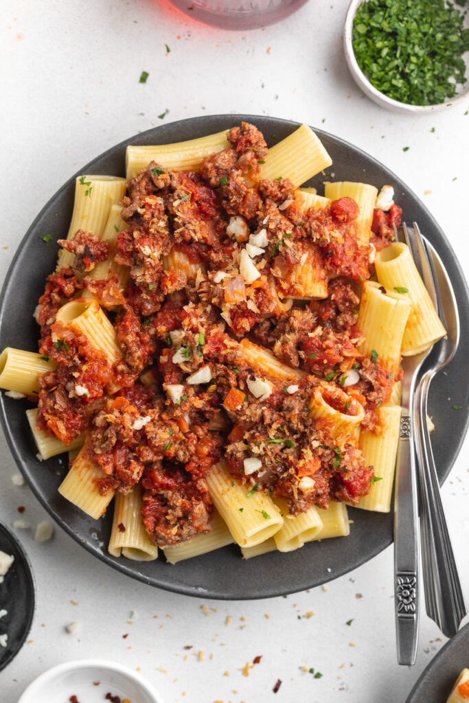 overhead image of rigatoni bolognese on a plate with red wine and chopped parsley on the side.
