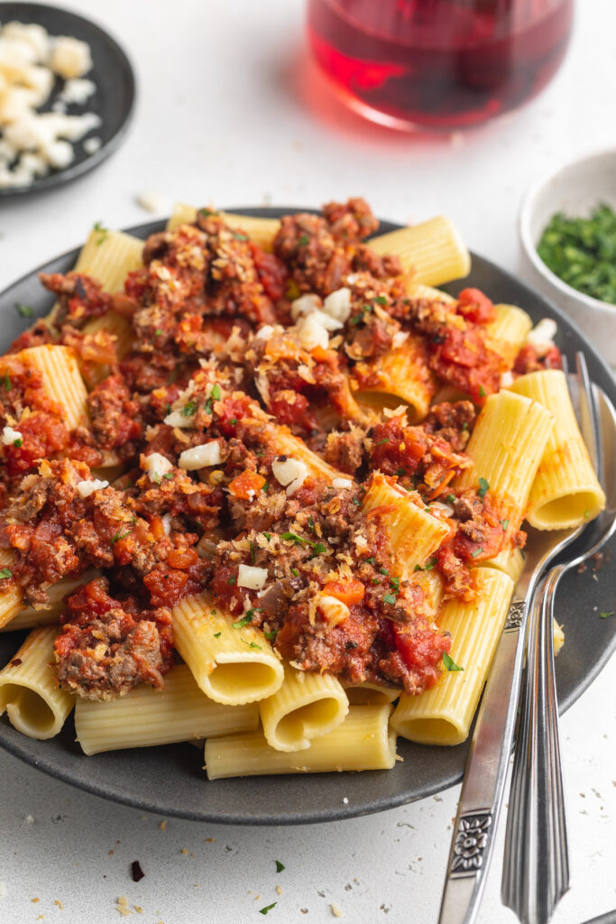 rigatoni bolognese on a plate with red wine and cheese on the side.